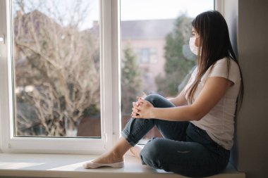 Pencerenin kenarında oturan ve koruyucu maskeli genç bir kadın. Pencereden dışarı bakıyor. Karantinada evde tek başına. Coronavirus teması. Evde kal.