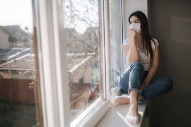 Pencerenin kenarında oturan ve koruyucu maskeli genç bir kadın. Pencereden dışarı bakıyor. Karantinada evde tek başına. Coronavirus teması. Evde kal.