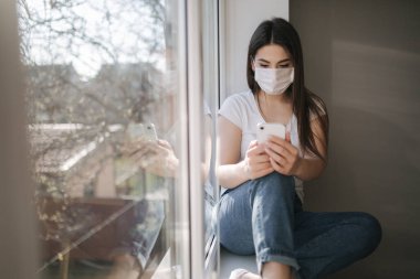 Tıbbi maskeli çekici bir kadın evdeki pencere eşiğinde oturuyor. Beyaz tişörtlü ve beyaz maskeli bir kadın telefon kullanıyor.