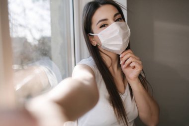 Tıbbi maskeli çekici kadın evde selfie çekiyor. Beyaz tişörtlü ve beyaz maskeli bir kadın telefon kullanıyor.