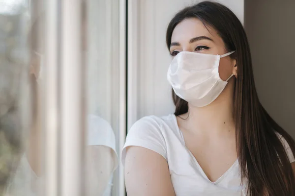 Genç bir kadın koruyucu maskeli pencere pervazında oturur ve pencereden dışarı bakar. Karantinada evde tek başına. Coronavirus teması. Evde kal. — Stok fotoğraf