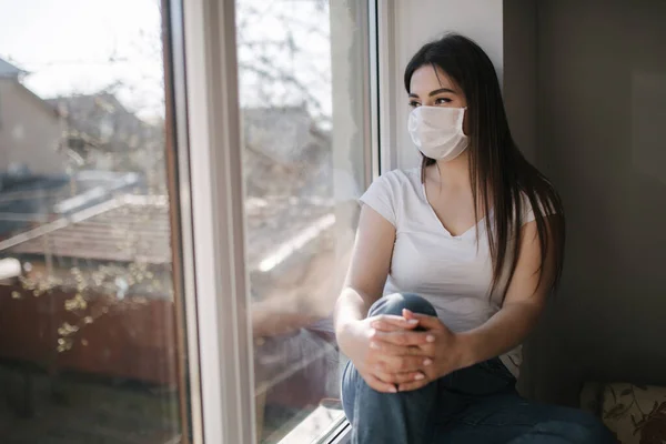 Widok z boku Młoda kobieta siedzi na parapecie i w masce ochronnej i wyjrzeć przez okno. Sam w domu, w kwarantannie. Motyw koronawirusa. Zostań w domu. — Zdjęcie stockowe