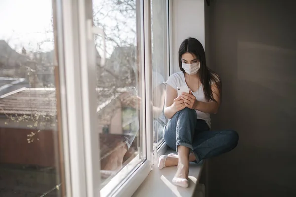 Vonzó nő orvosi maszkban ül az ablakpárkányon otthon. Nő fehér pólóban és fehér orvosi maszk használata telefon — Stock Fotó