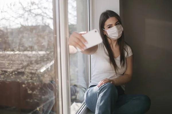 Tıbbi maskeli çekici kadın evde selfie çekiyor. Beyaz tişörtlü ve beyaz maskeli bir kadın telefon kullanıyor. — Stok fotoğraf