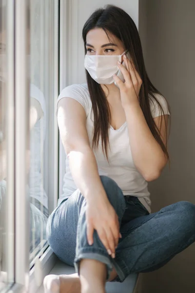 Tıbbi maskeli çekici bir kadın evdeki pencere eşiğinde oturuyor. Beyaz tişörtlü ve beyaz maskeli bir kadın telefon kullanıyor. — Stok fotoğraf