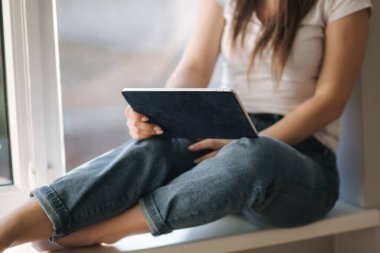 Beyaz tıbbi maskeli çekici genç kadının orta seçimi pencere kenarına oturup tablet kullanıyor. Kadın karantinada dinleniyor.