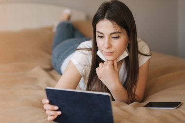 Genç kadın maskesini çıkar ve yatağa tablet koy. Karantina sırasındaki kadın videosu. Evde Öğrenci Çalışması