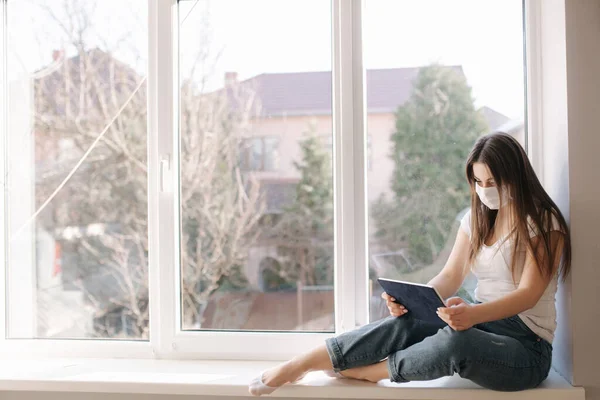 Привлекательная молодая женщина в белой медицинской маске сидит на подоконнике и пользуется планшетом. Женщины отдыхают дома в карантине — стоковое фото