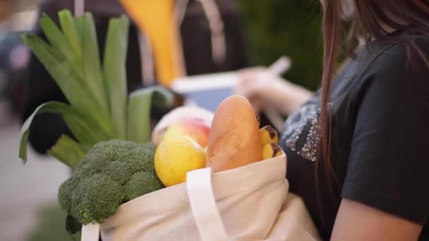 Serviço de entrega de comida inteligente. Homem em máscara médica e luvas entregando alimentos frescos para jovem cliente recebendo entrega expressa de correio em casa. Correio com tablet, iniciar sessão. Comida feminina — Vídeo de Stock