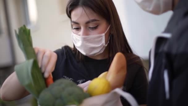 Servizio di consegna cibo intelligente. Uomo in maschera medica e guanti che consegna cibo fresco alla giovane cliente che riceve la consegna espressa dal corriere a casa. Corriere con tablet, accedi. Ordine femminile cibo — Video Stock