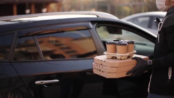 Kurir i skyddsmask och medicinska handskar tar order från bilen. Förlossningspojken håller pizza och kaffe. Leveransservice i karantän. Tema Coronavirus covid-19 — Stockvideo