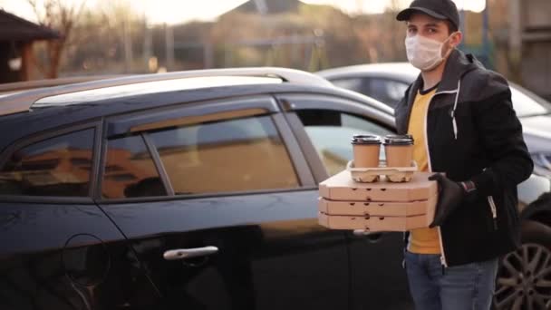 Kurir i skyddsmask och medicinska handskar tar order från bilen. Förlossningspojken håller pizza och kaffe. Leveransservice i karantän. Tema Coronavirus covid-19 — Stockvideo