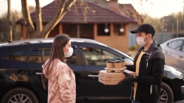 Jonge vrouw met beschermend masker en medisch masker neemt de bestelling van bezorger buiten. Voedsellevering in quarantaine. Courier brengen Pizza en koffie voor de klant — Stockvideo