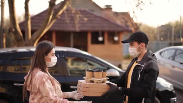 Νεαρή γυναίκα με προστατευτική μάσκα και ιατρική μάσκα παραγγέλνει από τον ντελιβερά έξω. Παράδοση τροφής σε καραντίνα. Courier φέρει πίτσα και καφέ για τον πελάτη — Αρχείο Βίντεο