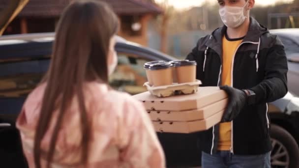 Jonge vrouw met beschermend masker en medisch masker neemt de bestelling van bezorger buiten. Voedsellevering in quarantaine. Courier brengen Pizza en koffie voor de klant — Stockvideo