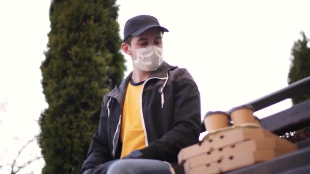 Kurir i skyddsmask och medicinska handskar levererar pizza och kaffe. Leverantörspojken sitter på bänken och ringer kunden. Leveransservice i karantän. Tema Coronavirus covid-19 — Stockvideo