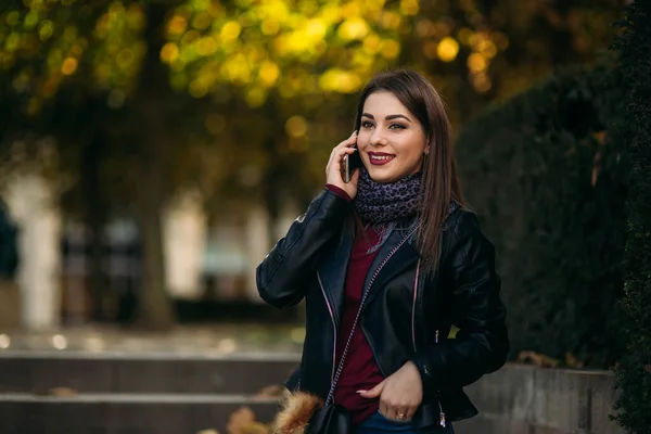 Stijlvolle mooie dame in een zwart leren jasje met een zwarte tas en bordeaux blouse. Aantrekkelijke jonge vrouw spreken door de telefoon — Stockfoto