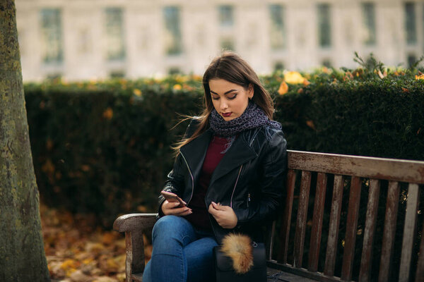 Stylish beautiful lady in a black leather jacket with a black bag and burgundy blouse. Attractive young woman use phone