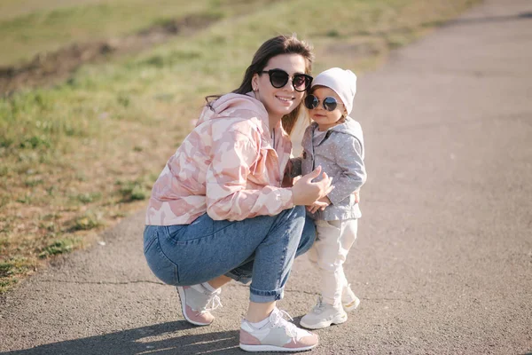 Glückliches kleines Mädchen geht mit ihrer Mutter im Park spazieren. Mutter und Tochter spazieren am Muttertag in der Nähe der Promenade. Nettes kleines Mädchen mit Sonnenbrille. Tochter umarmt Mama. Eineinhalbjähriges Mädchen — Stockfoto