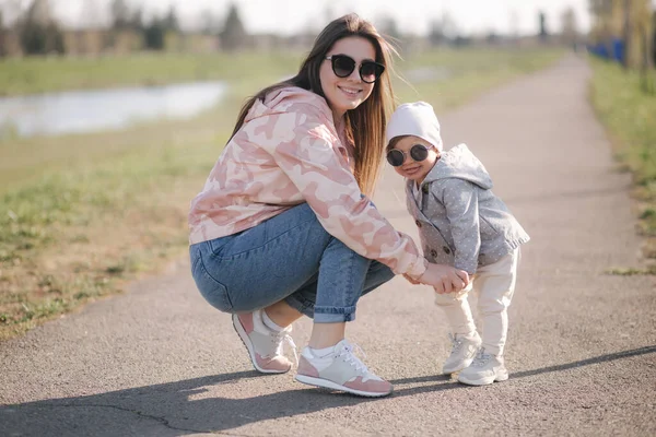 Šťastná holčička chodí se svou matkou do parku. Máma a dcera chodí v Den matek na promenádu. Roztomilá holčička ve slunečních brýlích. Dcera objímá mámu. Jedna a půl roku. — Stock fotografie