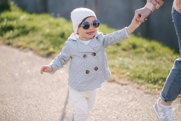 Boldog kislány sétál az anyjával a parkban. Anya és lánya sétálnak a sétányon az anyák napján. Cuki kislány napszemüvegben. — Stock Fotó