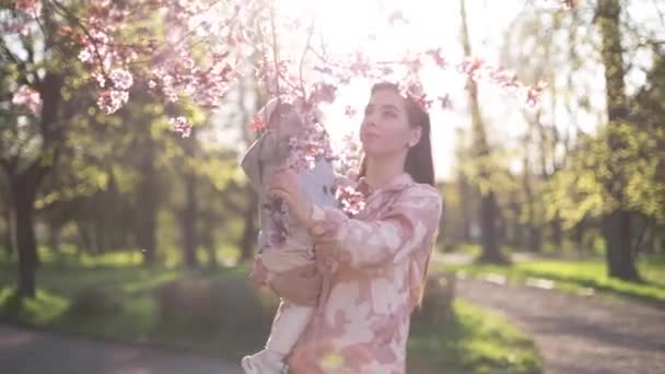 Krásná máma s rozkošnou dcerou stojí vedle růžového kvetoucího stromu. Šťastná rodina tráví čas na Den matek. Roztomilá holčička s matkou — Stock video