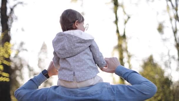 Urocza córeczka siedząca na szyi ojca i śmiejąca się. Młody ojciec spaceruje ze swoją uroczą córką w parku — Wideo stockowe
