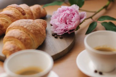 Kruvasanla kahve. Evdeki ahşap masada iki fincan kahve. Boş kruvasan. Masada pembe şakayık çiçeği