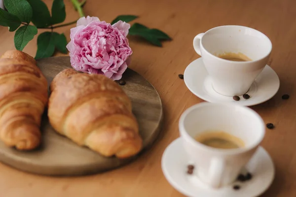 Kaffee mit Croissant vorhanden. Zwei Tassen Kaffee auf einem Holztisch zu Hause. Leeres Croissant. Rosa Pfingstrose Blume auf dem Tisch — Stockfoto