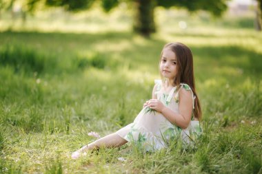 Sevimli küçük kızın portresi parktaki çimlerin üzerinde yalınayak oturuyor. Temiz havada mutlu bir çocuk. Karantina sona erdi. Güzel elbise.