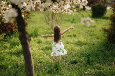 Yeşil çimenlerde yalın ayak yürüyen ve güzel çiçekli ağaçların etrafında dönen sevimli çocuk. Mutlu küçük kız, karantina sona erdi.