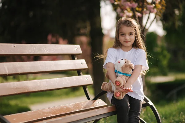 Happy female kid sits on the bench in the park. Fresh air. End of quarantine. Little girl hug teddy bear — Stock Photo, Image