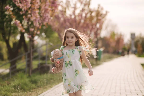 Маленька дівчинка гуляє в парку босоніж і обіймає плюшевого ведмедя — стокове фото