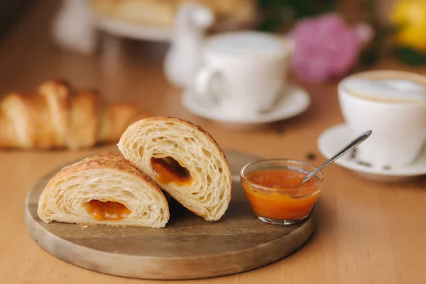 Zwei Hälften frisch entblößtes Croissant auf Holzbrett mit köstlicher hausgemachter Marillenmarmelade. Hintergrund des Cappuccino. Frühstück am Morgen — Stockfoto