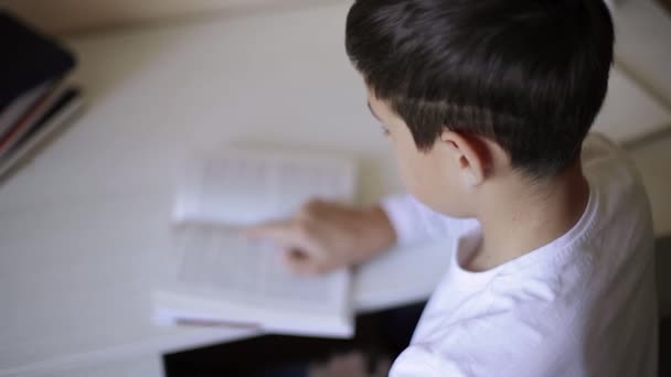 Młody chłopiec siedzący za biurkiem czyta książkę i zapisuje w notatniku. Ucz się w domu podczas kwarantanny. Kształcenie na odległość — Wideo stockowe