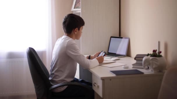 Schooljongen studeert aan een bureau in zijn kamer. Jongen gebruiken laptop en schrijven in notebook. Boeken en tablet op tafel. Onderzoek thuis tijdens qurantine — Stockvideo