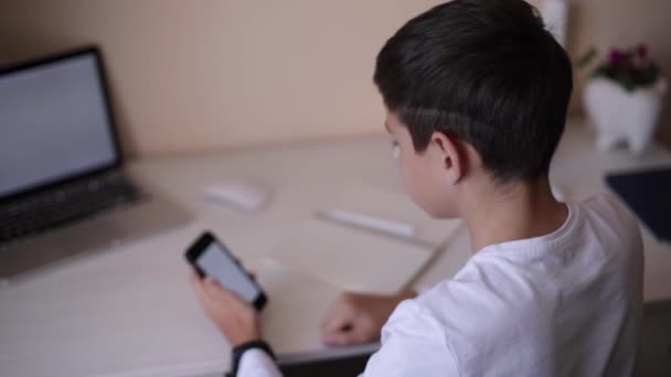 Skolpojke studerar vid skrivbordet i sitt rum. Pojken använder laptop och smartphone för att skriva i anteckningsboken. Böcker och surfplattor på bordet. Studera hemma under Qurantine — Stockvideo
