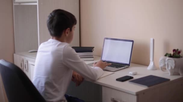 Estudiante de escuela en el escritorio de su habitación. El chico usa el portátil para las lecciones. Libros y tabletas sobre la mesa. Estudiar en casa durante la cuarentena — Vídeo de stock