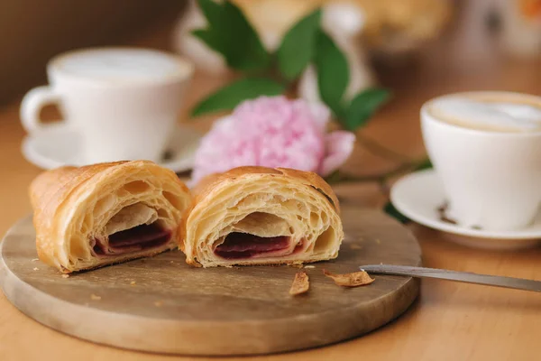 Zwei Hälften frisch entblößtes Croissant auf Holzbrett mit köstlicher hausgemachter Himbeermarmelade. Hintergrund des Cappuccino. Rosafarbene Pfingstrose — Stockfoto