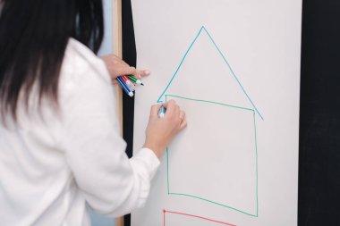 Öğretmen tahtaya yazıyor. Dişi bir ev çiziyor. Anaokulu