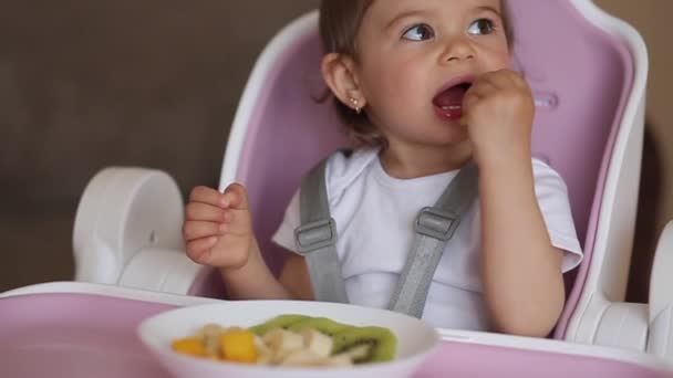 Vacker liten flicka första gången äta exotiska frukter i barnstol. Söt liten flicka smaka läcker fruktig, mango, kiwi och babana — Stockvideo
