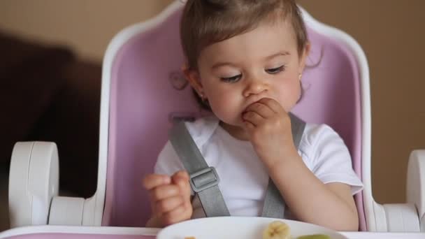 Linda menina primeira vez comendo frutas exóticas em cadeira alta. Menina bonito gosto delicioso fruist, manga, kiwi, e babana — Vídeo de Stock