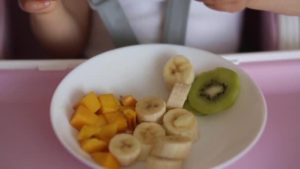 Close up de bebê menina tomar frutas exóticas da placa em cadeira alta. Manga cortada, kiwi e banana — Vídeo de Stock