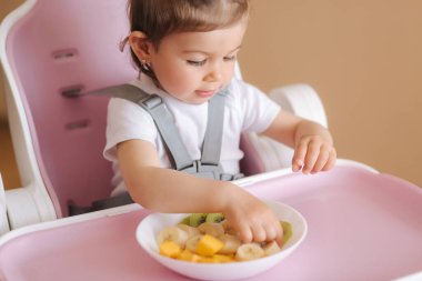 Güzel küçük kız ilk kez yüksek sandalyede egzotik meyveler yiyor. Tatlı bir kız bebek lezzetli bir düş kırıklığı, mango, kivi ve baban