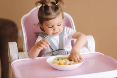 Güzel küçük kız ilk kez yüksek sandalyede egzotik meyveler yiyor. Tatlı bir kız bebek lezzetli bir düş kırıklığı, mango, kivi ve baban