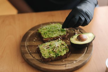 Çavdar ekmeği, guacamole ve mikro yeşillikle vejetaryen sandviç yapan bir kadın. Kadının üzerine susam tohumu serpiştirilmiş.