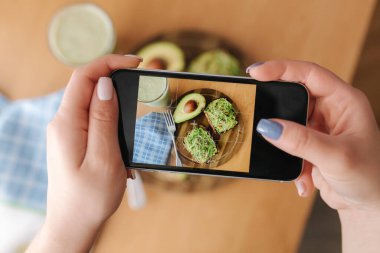 Kadın evde öğle yemeği fotoğrafı çekiyor. Dişi glogger yemek fotoğrafçılığı yapıyor. Sağlıklı vejetaryen yemeği. Avokadolu tost ve ıspanaklı smoothie.