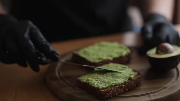 Close-up de Woman in black glowes coloca guacamole ou abacate espalhado em cima de pão de centeio torrada em tábua de madeira em casa. Café da manhã Vegan — Vídeo de Stock