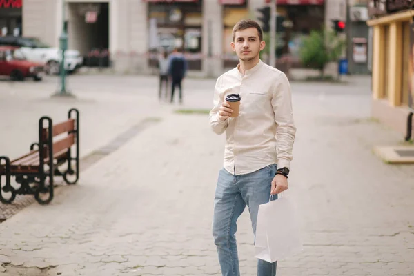 Şehirdeki yakışıklı genç adam, el işi paketi ve kahveyle ayakta duruyor. — Stok fotoğraf