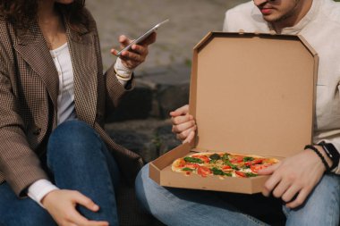 Genç blogcular açık havada vejetaryen pizzanın fotoğrafını çekiyor. Mutlu çift yemek fotoğrafçılığı yapar. Adam pizza kutusunu dizlerinin üstünde tutuyor. Vejetaryen fast food